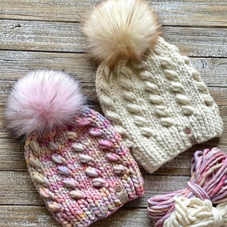Puede incluir: Dos gorros de punto con pompones esponjosos. Un gorro es blanco con un patr&oacute;n de punto de cable y el otro es de hilo multicolor con un patr&oacute;n de punto grueso. Ambos gorros tienen una etiqueta de cuero con el texto "The Wool Sheep" en ellos.
