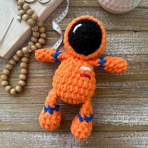 May include: A crochet plush toy shaped like an astronaut in a bright orange suit with a large black helmet visor, blue accents on the arms and legs, and small white and red rectangular details on the chest, crafted from soft yarn.