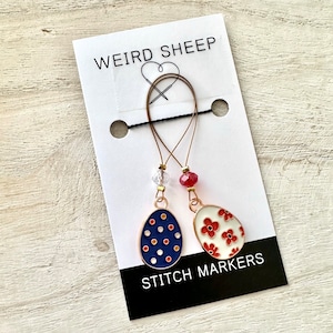 May include: Two gold stitch markers shaped like eggs. One marker has a blue and white polka dot design, and the other has a white background with red flowers. The markers are hanging from a card that says "Weird Sheep Stitch Markers".