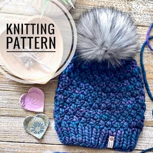 May include: A blue knitted beanie with a white pom pom and a small brown tag that says "Herb Sheep". The beanie is on a wooden surface with two heart-shaped dishes.