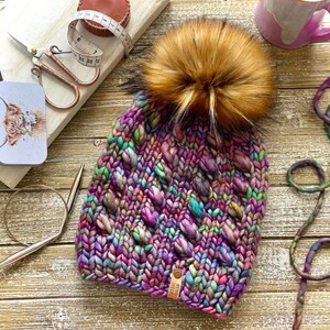 Puede incluir: Un gorro de punto colorido con un pomp&oacute;n de piel sint&eacute;tica. El gorro tiene una etiqueta de cuero marr&oacute;n con el texto "Weird Sheep" en &eacute;l.