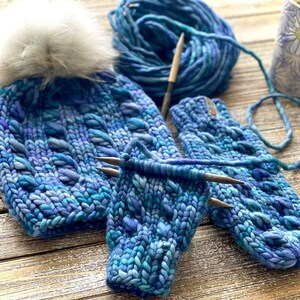 Puede incluir: Un gorro de punto azul y morado con un pomp&oacute;n blanco y guantes sin dedos a juego. El gorro y los guantes est&aacute;n hechos con un hilo grueso y tienen un patr&oacute;n de punto texturizado.