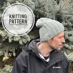 May include: A knitted hat in a light blue and white color with a ribbed texture. The hat is being worn by a person wearing a black jacket with a Columbia logo.