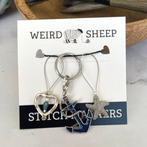 May include: Three silver stitch markers with charms. The charms are a heart with a blue bead, an anchor, and a starfish. The card they are attached to says "Weird Sheep Stitch Markers".