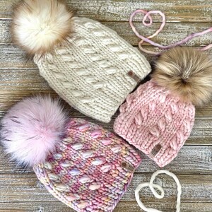 Puede incluir: Tres gorros de punto con pompones de piel sint&eacute;tica. Los gorros son en tonos de rosa y blanco. Los gorros tienen un patr&oacute;n de punto de cable y una etiqueta de cuero que dice "Knit With Love".