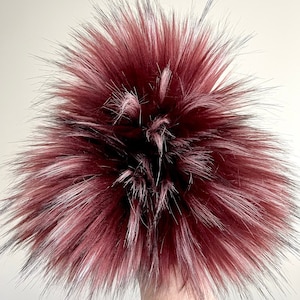 May include: A fluffy burgundy faux fur pom pom with black and white accents. The pom pom is made of soft, plush fur and is perfect for adding a touch of style to any hat or accessory.
