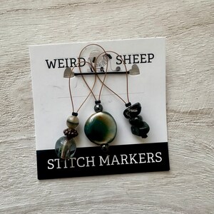 May include: Three stitch markers with different beads. The beads are black, green and brown. The stitch markers are labelled "Weird Sheep Stitch Markers".