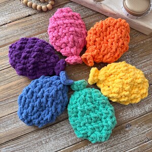 May include: Six handmade crochet water balloons in various colours: purple, pink, orange, blue, green, and yellow. The balloons are displayed on a wooden surface, highlighting their textured, plush design.