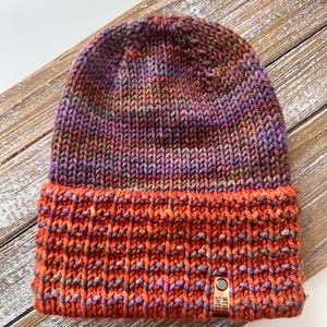 May include: A hand-knitted beanie hat in shades of orange, purple, and brown. The hat features a folded brim and a small leather tag that reads "WEIRD SHEEP". The hat is made of a thick, textured yarn.