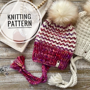 May include: A chunky knit beanie with a large faux fur pom-pom, featuring a chevron pattern in cream, magenta, and purple yarn. The hat includes braided earflaps with tassels and a small leather tag. Text reads KNITTING PATTERN.