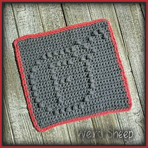 Puede incluir: Un paño de cocina de crochet gris con un diseño de corazón texturizado y un borde rojo. El paño de cocina está sobre una superficie de madera.