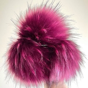 May include: A large, fluffy pom pom made of faux fur in a deep magenta colour. The pom pom is soft and plush, with a slightly textured appearance.