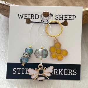 May include: Set of three stitch markers featuring a bee, honeycomb, and two crystal beads. The stitch markers are on a white card with the text "Weird Sheep Stitch Markers".