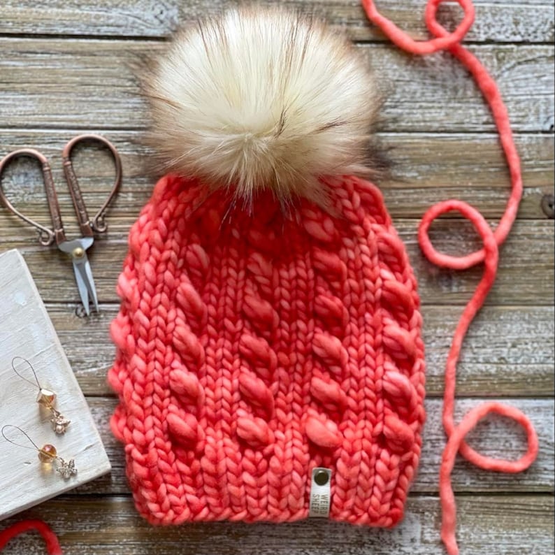 Puede incluir: Un gorro de punto de color coral con un pomp&oacute;n de piel sint&eacute;tica blanca. El gorro tiene un patr&oacute;n de punto de cable y una peque&ntilde;a etiqueta de cuero marr&oacute;n con texto.