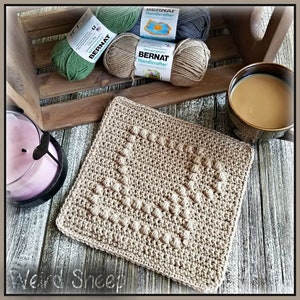 Könnte beinhalten: Ein quadratischer Häkel-Topflappen in beige mit einem strukturierten Muster. Der Topflappen liegt auf einer Holzoberfläche mit einer Kerze und Garnrollen im Hintergrund.