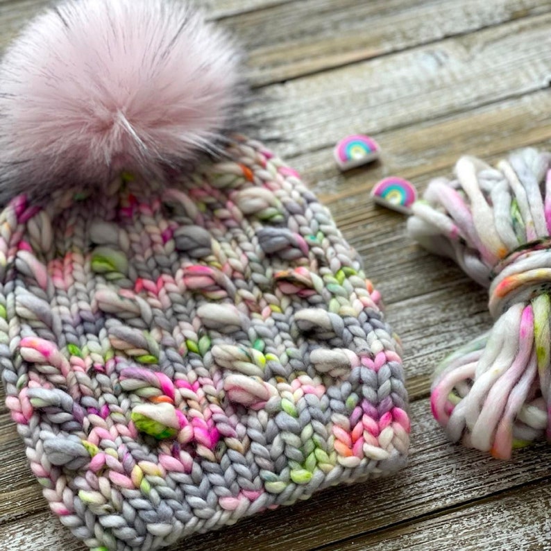 Puede incluir: Un gorro de punto gris, rosa y verde con un pomp&oacute;n rosa esponjoso. El gorro est&aacute; hecho con un hilo grueso y tiene un patr&oacute;n de punto texturizado. El hilo tambi&eacute;n se muestra en una madeja junto al gorro.