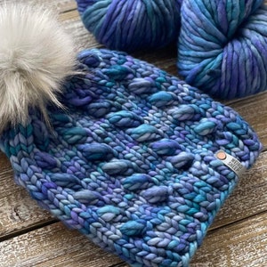 Puede incluir: Un gorro de punto azul y morado con un pomp&oacute;n blanco y una etiqueta "Weird Sheep". El gorro est&aacute; hecho con un hilo grueso y tiene un patr&oacute;n de punto de cable.
