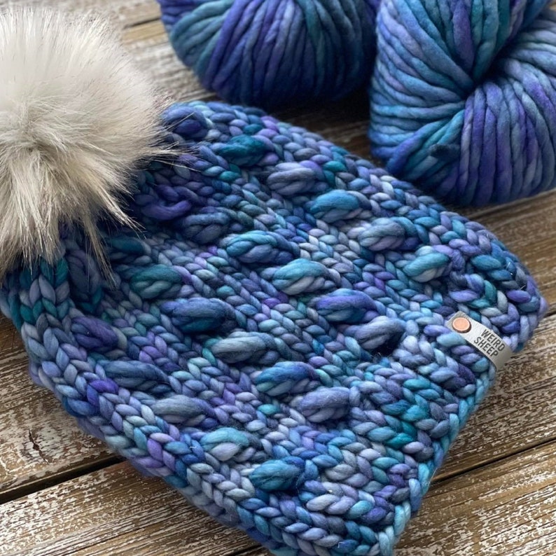 Puede incluir: Un gorro de punto azul y morado con un pomp&oacute;n blanco y una etiqueta "Weird Sheep". El gorro est&aacute; hecho con un hilo grueso y tiene un patr&oacute;n de punto de cable.
