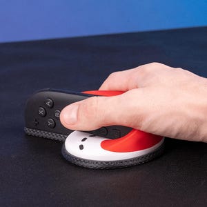Puede incluir: Un mando Joy-Con de Nintendo Switch negro y rojo junto a un agarre de mando blanco, rojo y negro. El agarre tiene un diseño curvo y está sujeto por una mano.