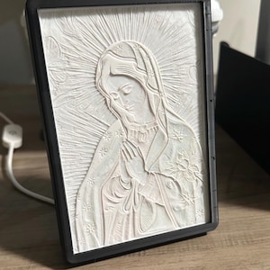 May include: A white relief sculpture of a religious figure, framed in black. The figure is depicted with hands clasped in prayer, wearing a veil and adorned with floral details. The background features radiating lines.