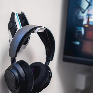 Puede incluir: Unos auriculares para juegos negros y grises colgados de un soporte de pared. El soporte es blanco, negro y gris con un acento azul. Los auriculares tienen grandes almohadillas negras con acolchado afelpado y una diadema gris.