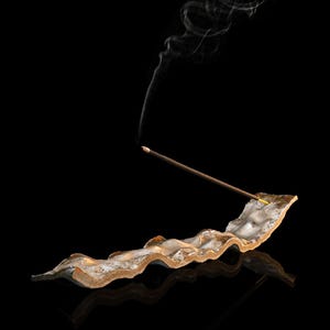 May include: A lit incense stick with wisps of smoke rising from a decorative, wavy, ceramic incense holder. The holder is a mix of beige, brown, and gold tones, set against a black background.