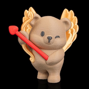 May include: A tan-colored bear figurine with gold-colored wings and a red bow and arrow. The bear is winking and has black eyes and a black nose. The figurine is standing on a black surface.
