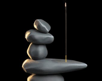 May include: A grey stone incense holder, featuring a stack of smooth, rounded stones. An incense stick is lit, with smoke rising against a black backdrop. The holder is designed to resemble a zen garden.