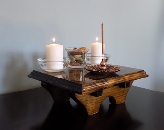 May include: A wooden altar with a dark brown stain and black trim. The altar has three legs and is decorated with two white candles, a small glass bowl filled with colorful stones, and a copper incense burner with a lit incense stick.