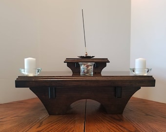 May include: A dark wood altar table with a raised platform. The table has a smooth, polished finish and features two small glass candle holders with white candles. A small glass jar with colorful stones sits on the platform, and a black incense stick is held in a metal holder above the jar.