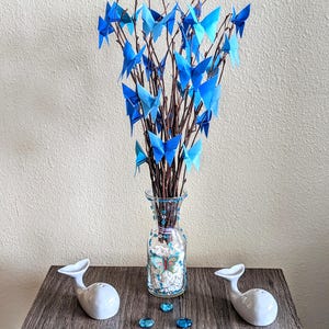 May include: A decorative arrangement featuring a glass vase filled with branches adorned with blue origami butterflies. Two white whale-shaped salt and pepper shakers sit on a wooden surface, along with blue decorative gems.