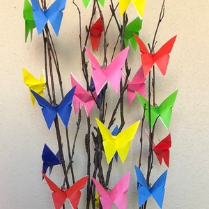 May include: A decorative arrangement featuring origami butterflies in various colors, including red, yellow, blue, and pink, attached to bare branches. The branches are placed in a black vase with a purple tissue paper base.