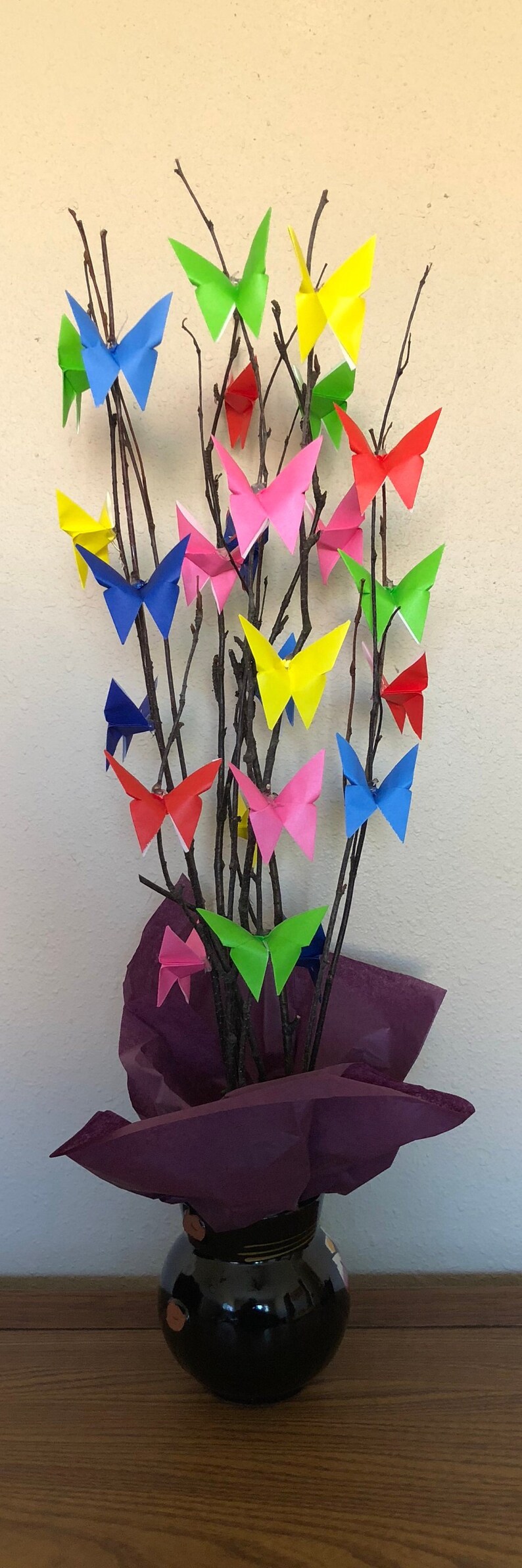 May include: A decorative arrangement featuring origami butterflies in various colors, including red, yellow, blue, and pink, attached to bare branches. The branches are placed in a black vase with a purple tissue paper base.