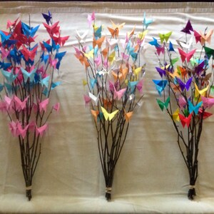 May include: Three bouquets of branches adorned with colorful origami butterflies. The butterflies are in shades of pink, blue, red, orange, yellow, green, and purple. The branches are tied together with twine.