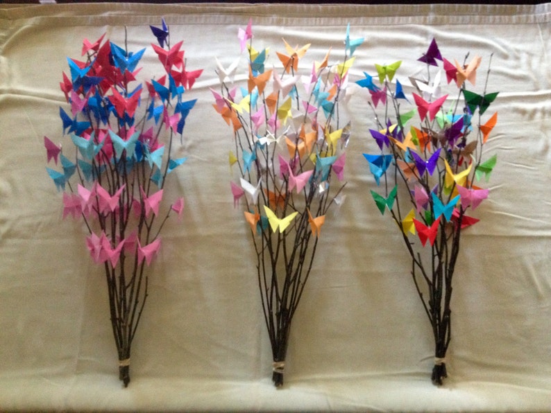 May include: Three bouquets of branches adorned with colorful origami butterflies. The butterflies are in shades of pink, blue, red, orange, yellow, green, and purple. The branches are tied together with twine.