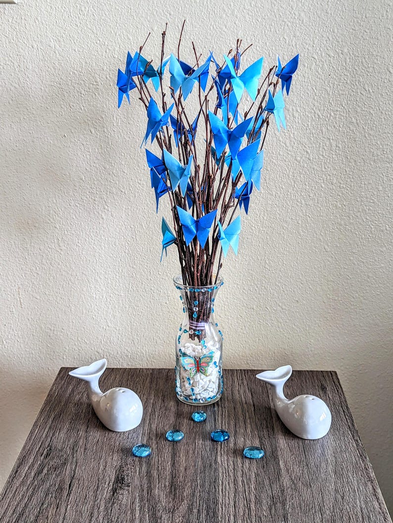 May include: A decorative arrangement featuring a glass vase filled with branches adorned with blue origami butterflies. Two white whale-shaped salt and pepper shakers sit on a wooden surface, along with blue decorative gems.