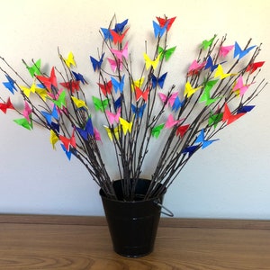 May include: A black bucket holds a collection of branches adorned with colorful origami butterflies. The butterflies are in shades of red, pink, yellow, blue, and green. The branches are dark brown, and the bucket has a silver handle.
