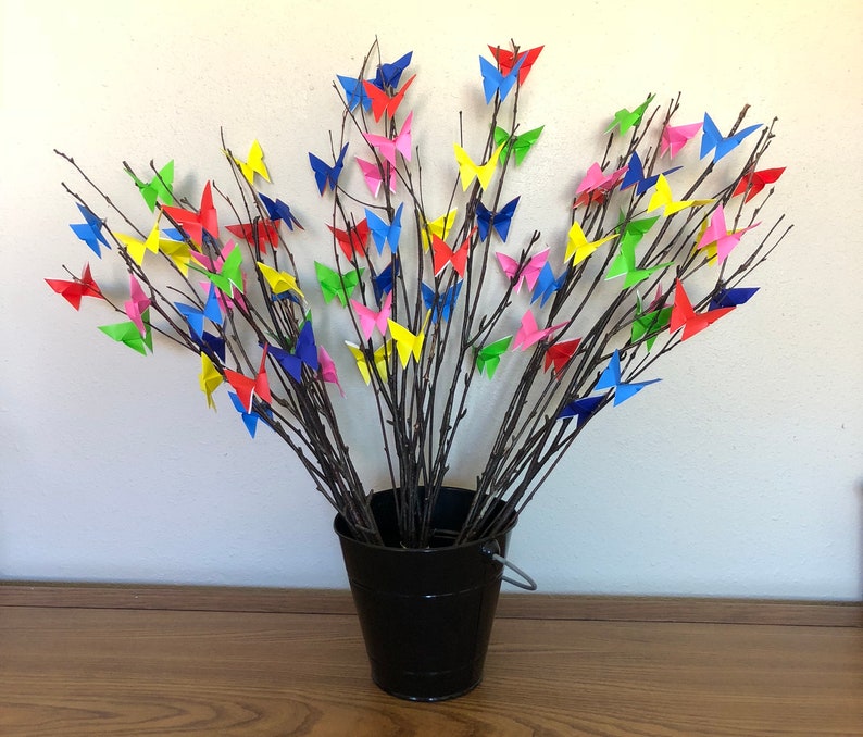 May include: A black bucket holds a collection of branches adorned with colorful origami butterflies. The butterflies are in shades of red, pink, yellow, blue, and green. The branches are dark brown, and the bucket has a silver handle.