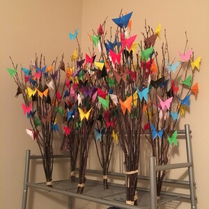 May include: Several bundles of tree branches adorned with colorful origami butterflies. The butterflies are in various colors, including blue, red, yellow, and green. The branches are tied together with twine and arranged on a metal shelf.