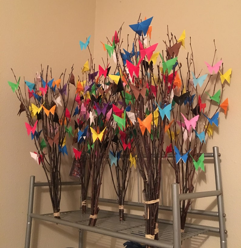 May include: Several bundles of tree branches adorned with colorful origami butterflies. The butterflies are in various colors, including blue, red, yellow, and green. The branches are tied together with twine and arranged on a metal shelf.