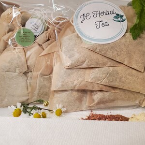 May include: A close-up of a package of loose leaf herbal tea bags. The tea bags are in a clear plastic bag with a white label that says "JE Herbal Tea". There are also some loose herbs and flowers scattered around the tea bags.