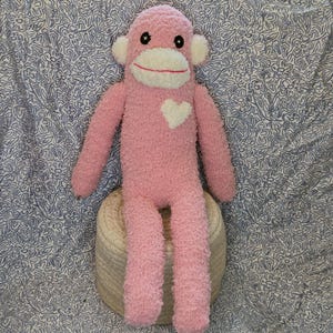 May include: A plush sock monkey toy made of fuzzy pink material with a white face, ears, and a heart-shaped patch on the chest. The monkey features black button eyes and a stitched red mouth, sitting upright with long arms and legs extended downward.