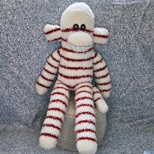 May include: A plush monkey toy crafted from fuzzy white fabric with horizontal dark red stripes. The toy features two round black button eyes, a stitched mouth, and long limbs. The texture of the material is soft and shaggy throughout the body.