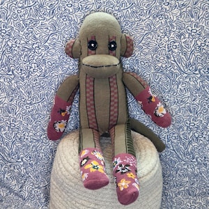 May include: A stuffed monkey toy crafted from olive green and brown striped fabric. It features black button eyes, a stitched mouth, and ears with a checkered pattern. The arms and legs are accented with a pink floral print fabric featuring yellow flowers.