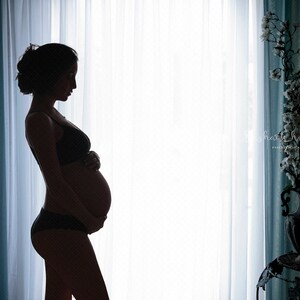 Könnte beinhalten: Silhouette einer schwangeren Person, die vor einem Fenster mit weißen Vorhängen steht. Die Person trägt einen BH und Unterwäsche. Das Fenster befindet sich hinter einem blauen Vorhang.
