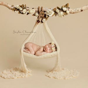 Könnte beinhalten: Ein neugeborenes Baby mit einer Blumenkrone schläft friedlich in einer weißen Stoffhängematte, die an einem mit weißen Blumen geschmückten Ast hängt.