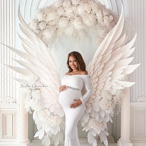 May include: A pregnant woman in a white off-the-shoulder gown with a sparkling belt stands in front of a white floral backdrop with large white angel wings.