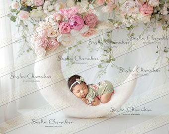 May include: A newborn baby girl sleeping in a white crescent moon shaped prop. The baby is wearing a green and pink floral onesie and a white headband with a pink bow. The background is white with a floral garland hanging above the baby.