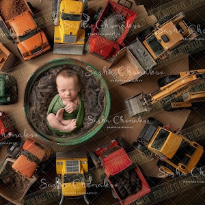 May include: A newborn baby boy sleeping in a green bucket surrounded by toy construction vehicles. The vehicles are yellow, red, green, and brown. The baby is wearing a green and white outfit.