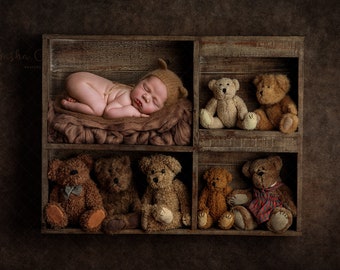 Könnte beinhalten: Ein neugeborenes Baby mit einer braunen Strickmütze schläft in einer Holzkiste mit mehreren braunen Teddybären. Die Kiste hat mehrere Regalbretter und steht vor einem braunen Hintergrund.